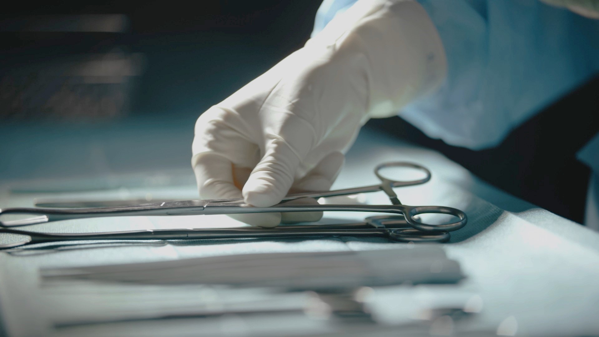 Bild einer Hand die eine Schere auf einem Tisch in einem OP-Saal aufhebt.
